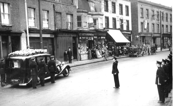 View of funeral car, with three uniformed firemen to each side and a senior officer saluting, with more firemen lining the roadside