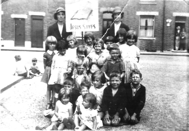 Group of around 20 girls and boys, ages from 3 or 4 to 10, with two lady teachers, placard 'Jesus Saves'