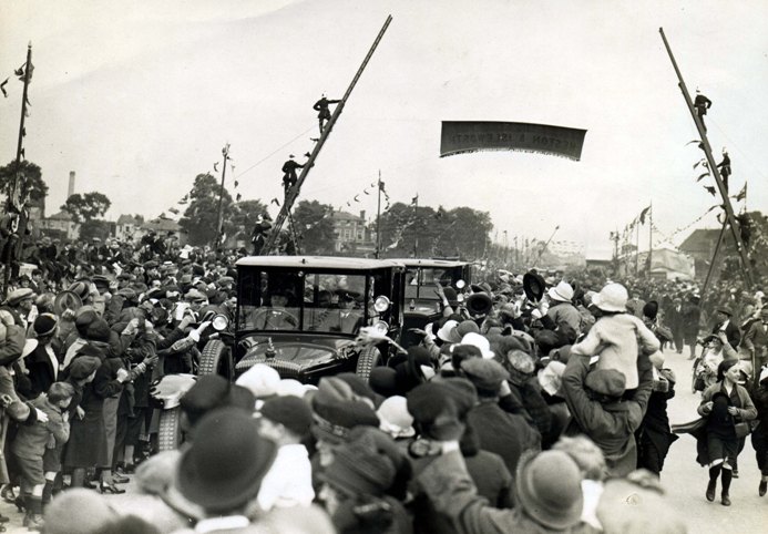 Great West Road lined with poles supporting bunting, two cars driving towards photographer surrounded by crowds of people